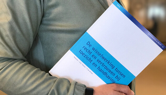 Persoon houdt boek 'De wisselwerking tussen recht en vertrouwen bij toezicht en handhaving' vast