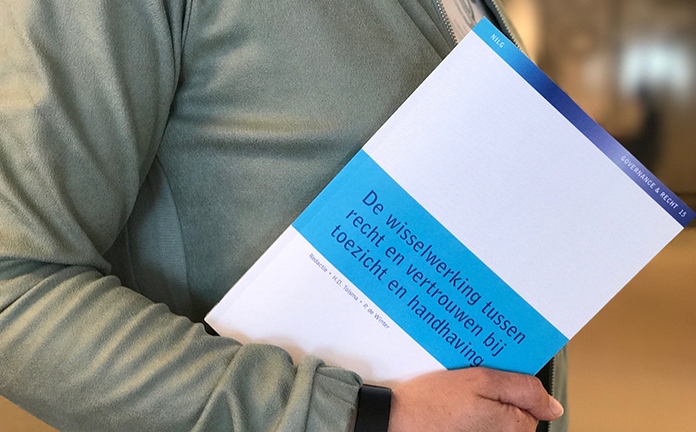 Persoon houdt boek 'De wisselwerking tussen recht en vertrouwen bij toezicht en handhaving' vast