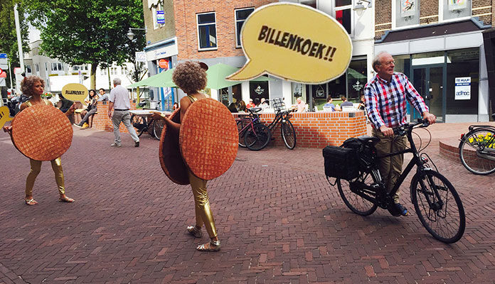 Mensen lopen op straat met de tekst 'Billenkoek' en grote stroopwafels.