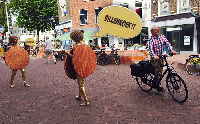 Mensen lopen op straat met de tekst 'Billenkoek' en grote stroopwafels.