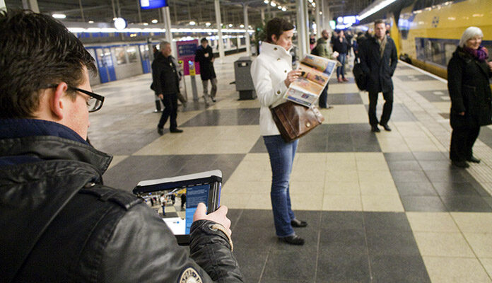 Een jonge man leest ToeZine op zijn Ipad terwijl hij wacht op de trein op treinstation Amsterdam Amstel. Ondertussen rijdt een trein het station in.