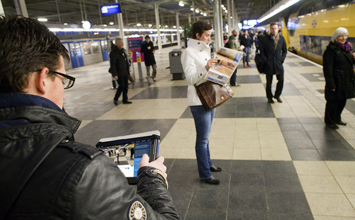 Een jonge man leest ToeZine op zijn Ipad terwijl hij wacht op de trein op treinstation Amsterdam Amstel. Ondertussen rijdt een trein het station in.
